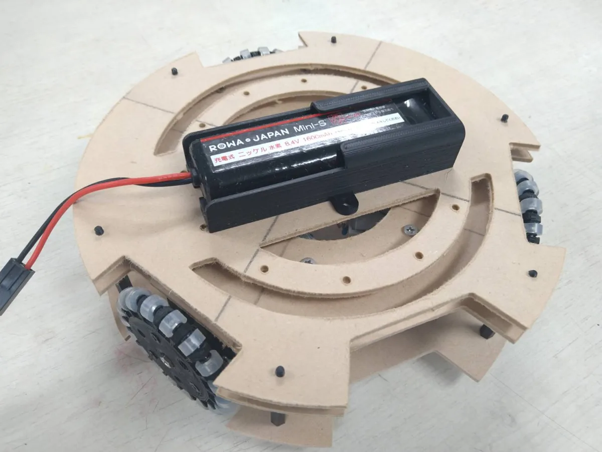 黒い樹脂で印刷した電池ボックスに 8.4V の充電池が入り、CNC で削った木の円形の土台の上に、耳のネジ穴で留められている。土台のまわりにはオムニホイールが付いている。