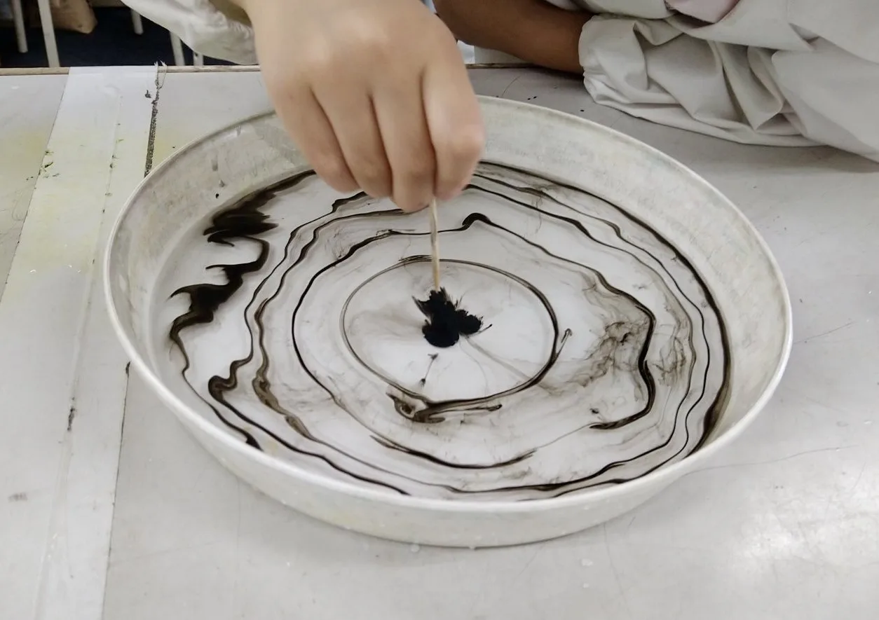 水を張ったバットの表面に墨が渦を巻いた模様を作っている様子。生徒が竹串で墨に触れている